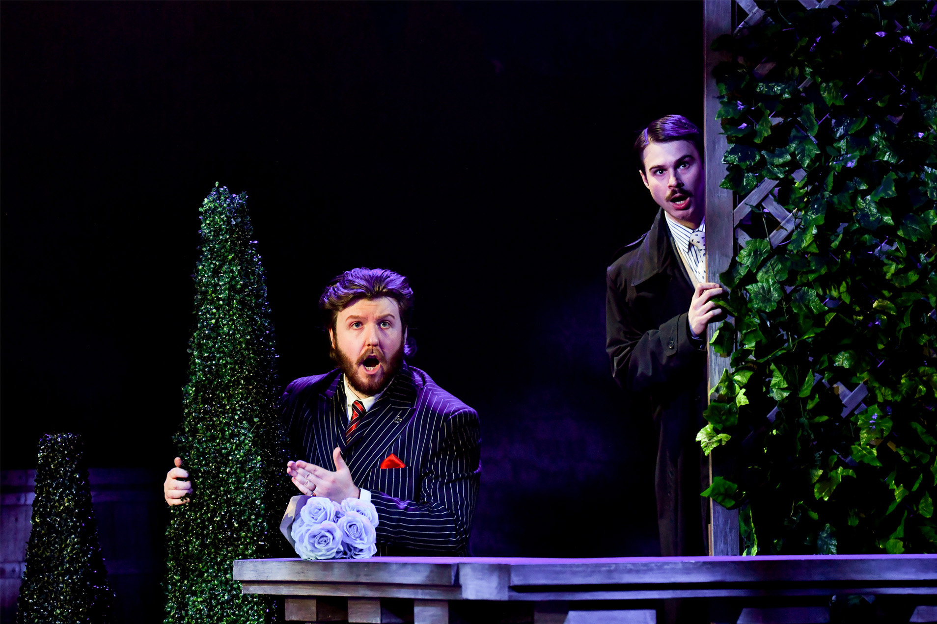 Two male opera singers performing on stage, hiding and singing behind shrubs.